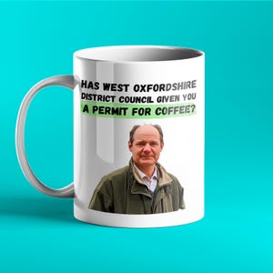 Op de afbeelding: Witte keramische mok met een handvat. De mok toont de tekst "HAS WEST OXFORDSHIRE DISTRICT COUNCIL GIVEN YOU A PERMIT FOR COFFEE?" en een portret van een man. De mok staat tegen een turquoise achtergrond.