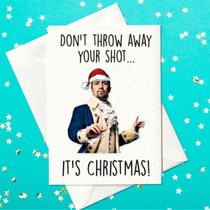 May include: A white Christmas card with the text "DON'T THROW AWAY YOUR SHOT... IT'S CHRISTMAS!". The card features a man in a Santa hat and historical attire. The card is inside a white envelope, set against a teal background with star-shaped confetti.