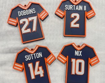 May include: Four navy blue jersey ornaments with orange and white accents. Each ornament features a different name and number: Dobbins 27, Surtain II 2, Sutton 14, and Nix 10. The ornaments are shaped like football jerseys.