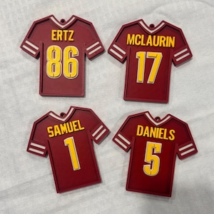 May include: Four maroon jersey-shaped ornaments with white stripes and yellow numbers and names. The jerseys are labeled: ERTZ 86, MCLAURIN 17, SAMUEL 1, and DANIELS 5. Each ornament has a small hole at the top.