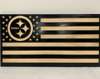 May include: A wooden American flag with a black background and natural wood stripes. The flag features the Pittsburgh Steelers logo in the upper left corner.