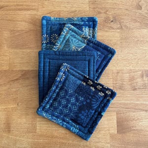 May include: A set of four square fabric coasters featuring various shades of blue and indigo. Each coaster displays a unique geometric or floral pattern with intricate white and light blue details, finished with quilted stitching along the edges.