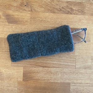 May include: A gray felt glasses case with a pair of eyeglasses partially inside. The case is rectangular and has a soft, textured appearance. The eyeglasses have a dark frame and clear lenses.