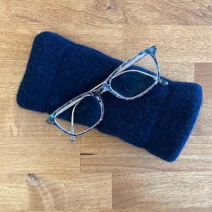 May include: A pair of eyeglasses with a speckled pattern frame rests on a dark blue, rectangular glasses case. The glasses have clear lenses and thin metal arms. The case is made of a soft, knitted material.