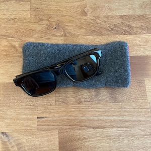 May include: Black sunglasses with a rectangular frame rest on a gray felt case. The sunglasses have dark lenses and a sleek, modern design. The case is rectangular and provides protection for the eyewear.