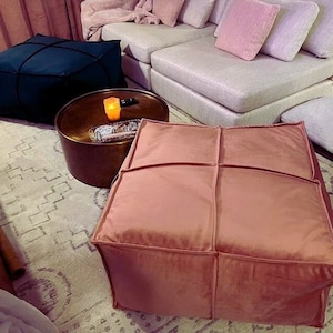 May include: A blush pink velvet ottoman with a square shape and four stitched panels. The ottoman is sitting on a rug with a geometric pattern.