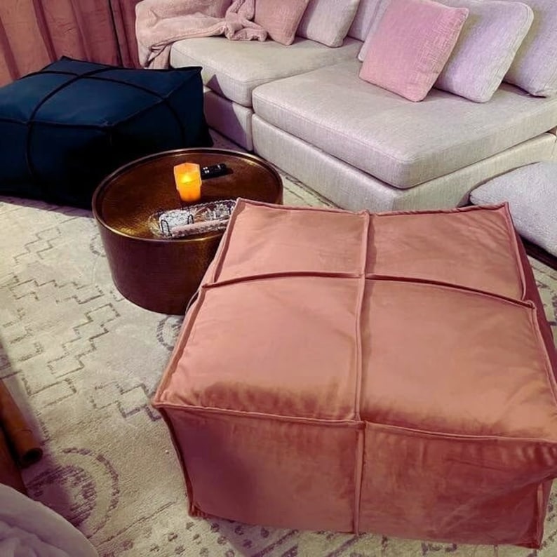 May include: A blush pink velvet ottoman with a square shape and four stitched panels. The ottoman is sitting on a rug with a geometric pattern.