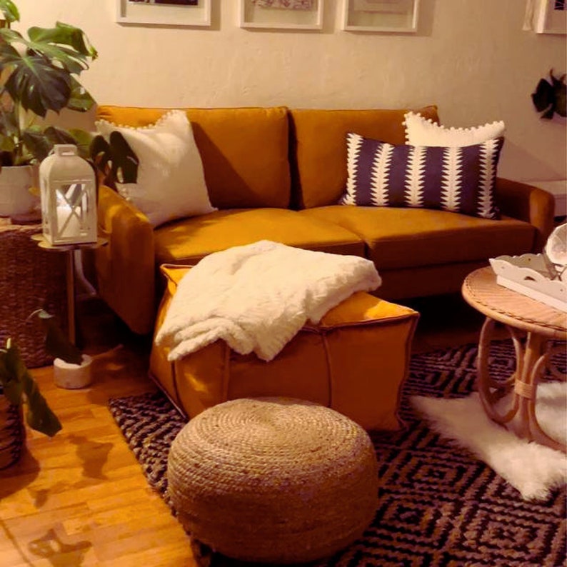 May include: A mustard yellow sofa with white and black patterned pillows, a white throw blanket on a mustard yellow ottoman, and a woven jute footstool. The sofa is in a living room with a wooden floor and a patterned rug.