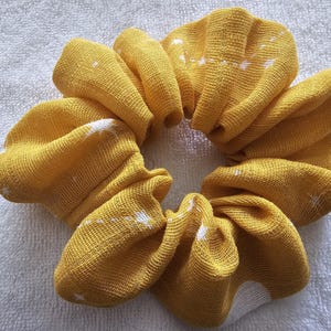 May include: A yellow scrunchie with a white star pattern. The fabric is gathered and folded, creating a textured, circular shape. The scrunchie is made of a soft material and is suitable for various hairstyles.