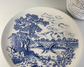 May include: A white ceramic plate with a blue landscape design. The design features a bridge over a river, trees, mountains, and clouds. The plate has a classic, vintage aesthetic.
