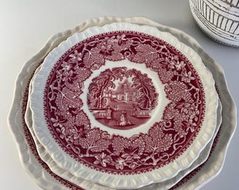 May include: Three white ceramic plates with a scalloped edge. The plates are stacked on top of each other. The top plate has a red and white floral design with a central image of a building and trees.