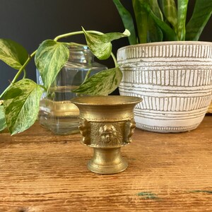 May include: A small, gold-colored metal decorative urn with three faces on the side. The urn is sitting on a wooden surface.