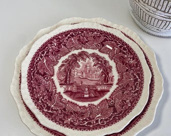May include: Two vintage pink and white ceramic plates with a scalloped edge. The top plate features a detailed floral design with a central image of a building and trees.