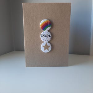 May include: A brown card with three round buttons attached. The top button has a rainbow design. The middle button has the word "Dioleh" on it. The bottom button has a gold star on it.