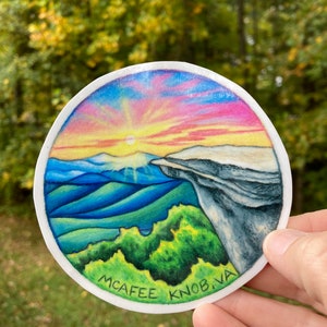 May include: A round sticker with a colorful illustration of a mountain range with a sunset sky. The sticker has the text "McAfee Knob, VA" at the bottom.