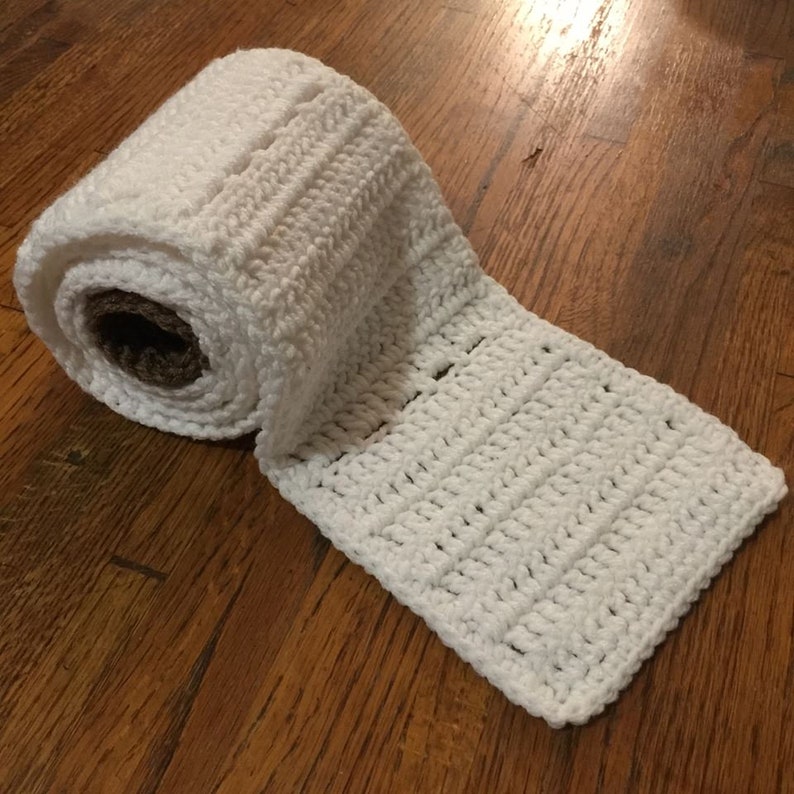 Może przedstawiać: Biały, szydełkowy rulon papieru toaletowego z brązowym kartonowym środkiem. Szydełkowy rulon jest rozwinięty i leży płasko na drewnianej powierzchni.