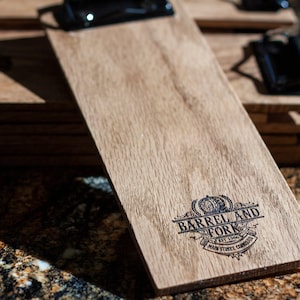 May include: A wooden menu board with a black metal clip. The board is engraved with the text "Barrel and Fork" and the logo includes a barrel and fork design. The logo also includes the text "Est. 1996" and "Main Street, Colorado".