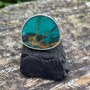 May include: A ring featuring a large, rounded, teal-colored stone with prominent brown and gold veining. The stone is set in a smooth, polished silver-toned metal bezel that follows the irregular, organic shape of the gemstone's perimeter.