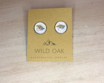 May include: A pair of round, white, resin earrings with a colorful fly fishing lure design. The earrings are displayed on a brown card with the text "WILD OAK HANDCRAFTED JEWELRY".