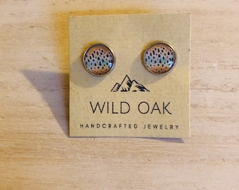 May include: A pair of silver stud earrings with a round, colorful design. The design features a brown, blue, and white pattern that resembles a trout fish. The earrings are displayed on a card with the text "WILD OAK HANDCRAFTED JEWELLERY".