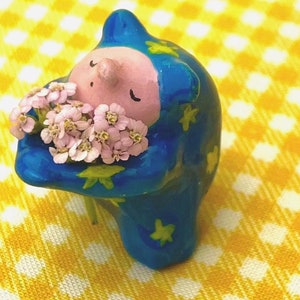 May include: A blue ceramic figurine with a pink face and a bouquet of pink flowers in its arms. The figurine is standing on a yellow and white checkered surface.