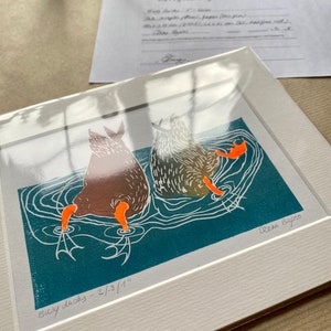 Puede incluir: Una impresión enmarcada de dos patos nadando en un estanque azul verdoso. La impresión está titulada "Busy Ducks - 2/8/1" y firmada "Elena Bagno".