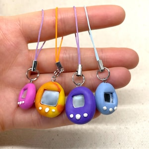 May include: Five colorful, egg-shaped, keychain charms with a screen and buttons, resembling a popular handheld game. Each charm has a different color: pink, orange, purple, blue, and light blue. The charms are attached to colorful strings.