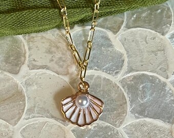 May include: Gold-toned necklace featuring a seashell pendant with a pearl center. The pendant has a pink and white enamel design. The chain is a delicate link style, and the background includes a green fabric and a textured surface.