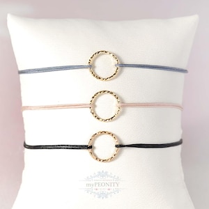 Op de afbeelding: Drie minimalistische armbanden met goudkleurige, getextureerde cirkelbedels. Elke armband heeft een verschillend gekleurd koord: grijs, roze en zwart. De armbanden worden gepresenteerd op een wit kussen tegen een zachtroze achtergrond.