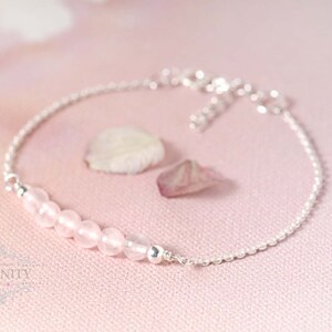 Puede incluir: Un delicado brazalete de cadena de plata con una fila de cuentas de piedras preciosas de color rosa claro. El brazalete est&aacute; sobre un fondo rosa.