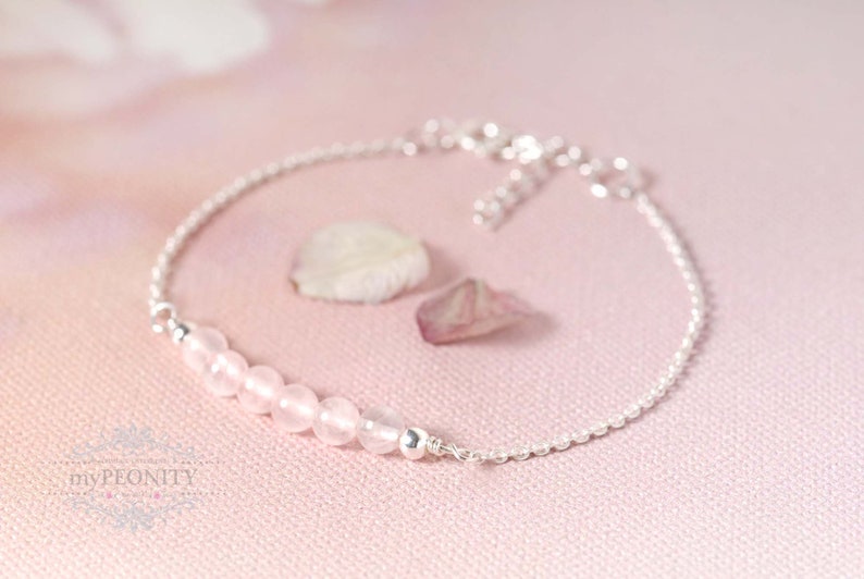 Puede incluir: Un delicado brazalete de cadena de plata con una fila de cuentas de piedras preciosas de color rosa claro. El brazalete est&aacute; sobre un fondo rosa.