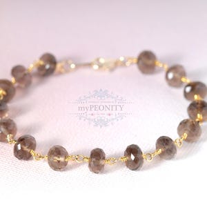 Könnte beinhalten: Ein zartes Armband aus goldener Kette mit Rauchquarzperlen. Das Armband ist in einem schlichten Design gehalten und verfügt über einen Verschluss.