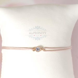 Könnte beinhalten: Ein zartes rosafarbenes Armband aus Schnur mit einem goldfarbenen Verschluss und einem klaren Kristall-Akzent. Das Armband wird auf einem weißen Kissen präsentiert.