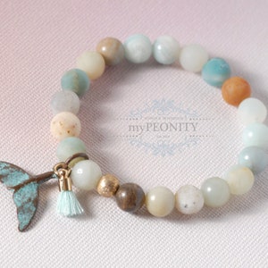 Op de afbeelding: Een lichtblauwe, groene en bruine kralen armband met een turquoise zeemeerminstaart bedel en een lichtblauwe kwast. De armband ligt op een witte, textuur achtergrond.