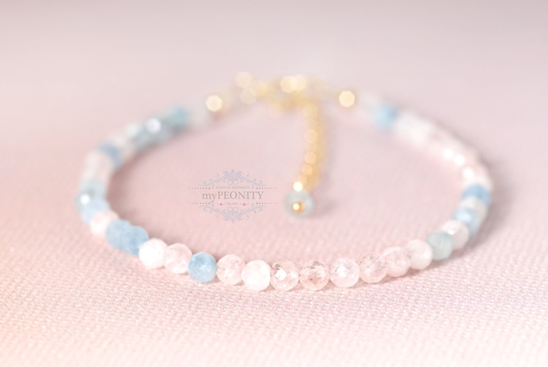 May include: A delicate bracelet made with light blue and pink gemstone beads. The bracelet has a gold clasp and is strung on a clear elastic cord.