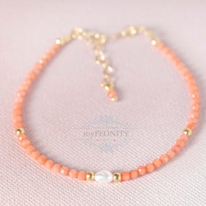 Op de afbeelding: Een delicate armband gemaakt van kleine, lichtroze koraal kralen en een enkele witte parel. De armband heeft een gouden sluiting en kleine gouden kralen die door de hele armband verspreid zijn.