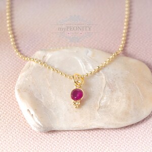 Pode incluir: Um colar de corrente de contas douradas com um pequeno pingente de pedra preciosa rosa-escuro, engastado numa moldura dourada com um minúsculo detalhe de conta dourada na base. O pingente está suspenso numa delicada corrente dourada.