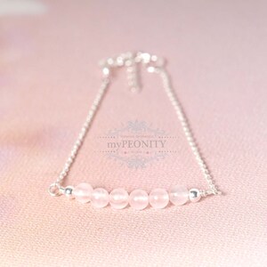 Puede incluir: Un delicado brazalete de cadena de plata con una fila de seis cuentas de color rosa p&aacute;lido. El brazalete est&aacute; sobre un fondo rosa.