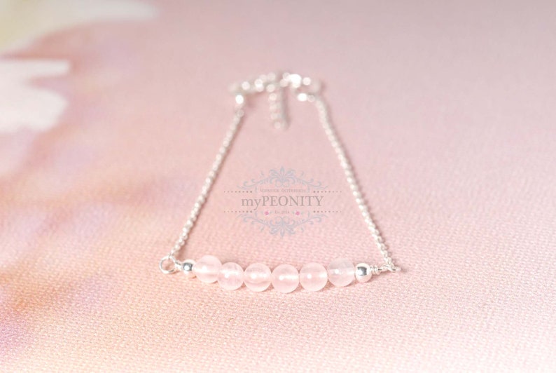 Puede incluir: Un delicado brazalete de cadena de plata con una fila de seis cuentas de color rosa p&aacute;lido. El brazalete est&aacute; sobre un fondo rosa.