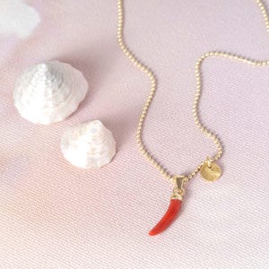 Könnte beinhalten: Eine goldfarbene Perlenketten-Halskette mit einem kleinen, gebogenen, hornförmigen roten Anhänger und einem winzigen runden Goldanhänger. Die Kette liegt neben zwei weißen, spiralförmigen Muscheln auf einer hellen, strukturierten Oberfläche.