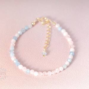 May include: A delicate bracelet made with light blue and pink gemstone beads. The bracelet has a gold clasp and is strung on a clear elastic cord.