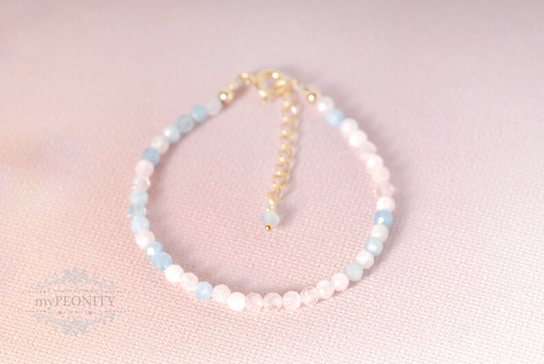 May include: A delicate bracelet made with light blue and pink gemstone beads. The bracelet has a gold clasp and is strung on a clear elastic cord.