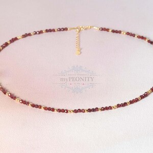 Op de afbeelding: Fijne ketting met kleine, dieprode edelstenen en goudkleurige kralen. De ketting is geregen op een fijne ketting en heeft een verstelbare lengte. Het ontwerp is eenvoudig en elegant, geschikt voor dagelijks gebruik of speciale gelegenheden.