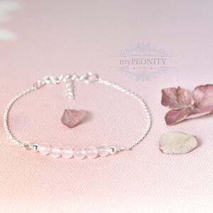 Puede incluir: Un delicado brazalete de cadena de plata con una fila de cuentas de cuarzo rosa p&aacute;lido. El brazalete est&aacute; sobre un fondo rosa con p&eacute;talos de flores secas.