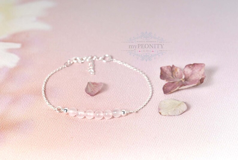 Puede incluir: Un delicado brazalete de cadena de plata con una fila de cuentas de cuarzo rosa p&aacute;lido. El brazalete est&aacute; sobre un fondo rosa con p&eacute;talos de flores secas.