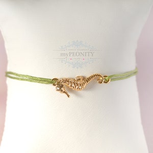 Op de afbeelding: Een delicate armband met een goudkleurige zeepaardje-bedel in het midden van een dubbelstrengs lichtgroen koord. De bedel heeft een gestructureerd, gedetailleerd zeepaardje-ontwerp. Boven de armband staat de tekst myPEONITY in een decoratief lettertype.