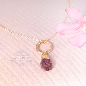 Op de afbeelding: Een gouden ketting met een delicate ronde hanger en een hangende roze edelsteen. De ketting wordt getoond op een roze achtergrond met een roze bloem.