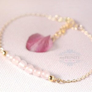 Puede incluir: Un delicado brazalete de cadena dorada con una hebra de cuentas de cuarzo rosa y un dije de flor rosa. El brazalete est&aacute; sobre un fondo blanco.