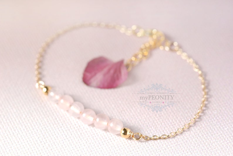 Puede incluir: Un delicado brazalete de cadena dorada con una hebra de cuentas de cuarzo rosa y un dije de flor rosa. El brazalete est&aacute; sobre un fondo blanco.