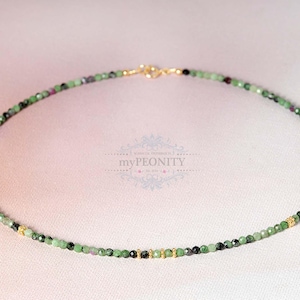 Peut inclure: Un collier délicat en pierres précieuses vertes et noires avec des accents dorés. Le collier est composé de petites perles rondes et est enfilé sur une fine chaîne.